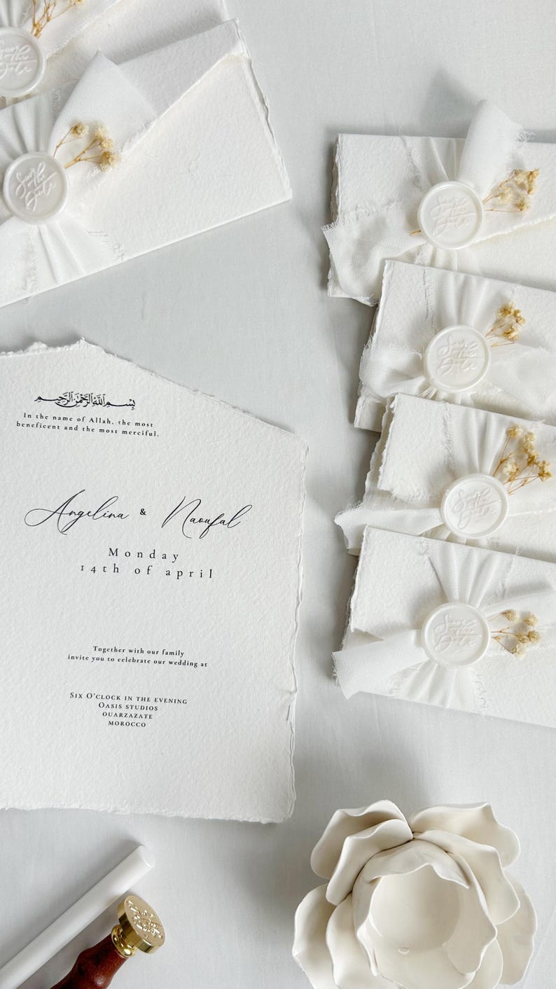 Op de afbeelding: Een witte trouwkaart met gescheurde randen en zwarte tekst met de namen Angelina en Naufal, samen met bijpassende witte enveloppen verzegeld met witte waszegels, wit lint en kleine gedroogde bloemen.