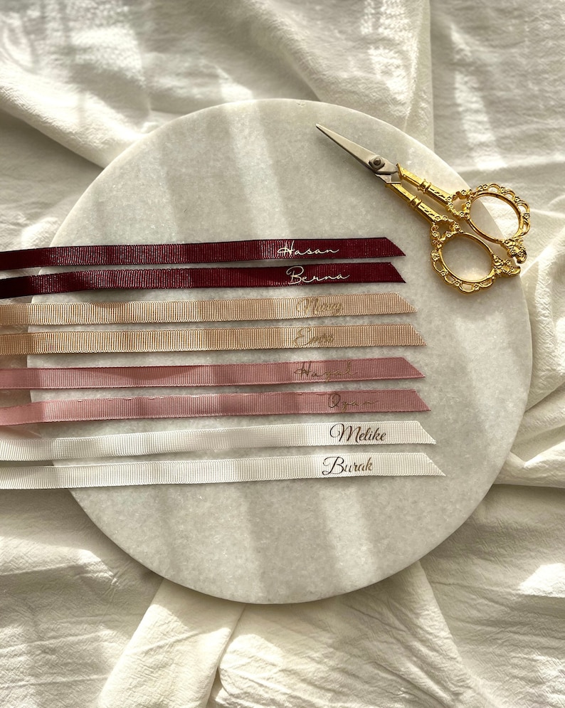 May include: Six satin ribbons with gold lettering on a white marble surface. The ribbons are in shades of burgundy, beige, and pink. The names on the ribbons are Hasan, Berna, Melisa, Elif, Hamza, Ozgur, Mellike, and Burak.