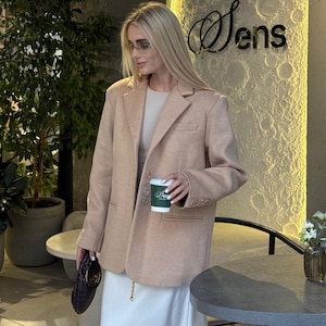 May include: A tan blazer with a classic lapel and button closure. The blazer is paired with a white dress and a small black handbag. The image also includes a coffee cup with a green and white design.