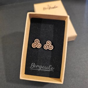Könnte beinhalten: Holz-Ohrstecker mit keltischem Spiralmuster, präsentiert in einer kleinen, offenen Pappschachtel mit schwarzem Innenraum. Die Ohrringe sind hellbraun, und die Schachtel trägt den Markennamen "Bergeule".