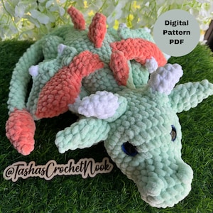 Könnte beinhalten: Ein grüner Häkel-Drache mit rosa und weißen Akzenten. Der Drache liegt auf der Seite mit dem Kopf nach rechts gedreht. Der Text "Digital Pattern PDF" und "@TashasCrochetNook" ist auf dem Bild zu sehen.