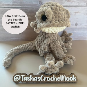 以下が含まれることがあります： ベージュに白いアクセントがついた、かぎ針編みで作られたヒゲを生やしたドラゴンぬいぐるみ。ぬいぐるみは木製の表面に置かれ、「@TashasCrochetNook」と書かれた看板がある。画像には「LOW SEW-Beau the Beardie PATTERN-PDF-English」というテキストが表示されている。