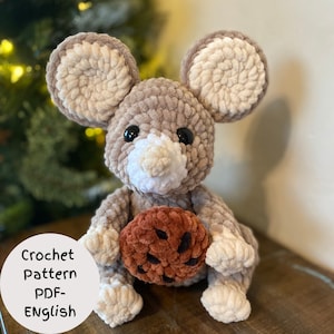 May include: A crocheted plush mouse toy holding a brown crocheted cookie. The mouse is beige with white accents and black eyes. The toy is sitting on a wooden surface. Crochet Pattern PDF - English
