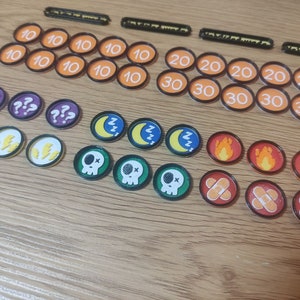 Puede incluir: Una colección de fichas de juego coloridas con varios símbolos y números. Las fichas incluyen signos de interrogación, rayos, lunas durmientes, calaveras, llamas y vendas. También hay fichas naranjas y rojas con los números 10, 20 y 30.