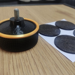 Op de afbeelding: Een zwarte en oranje meubelpoten met een metalen schroefdraadstang en een getextureerde basis. Daarnaast liggen verschillende donkergrijze, ovale viltjes op een wit vel papier. De poot is ontworpen om aan meubels te worden bevestigd.