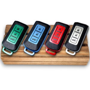 Op de afbeelding: Vier autosleutelhangers in verschillende kleuren: groen, blauw, rood en zilver. Elke sleutelhanger heeft een zwarte, getextureerde hoes en een gekleurd frame en handvat. De sleutels zijn gerangschikt op een houten oppervlak.