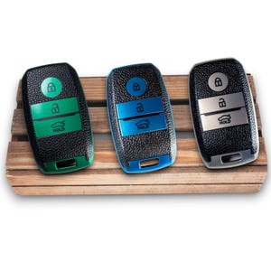Peut inclure: Trois télécommandes de voiture avec des coques noires texturées et des accents colorés. L'une a des accents verts, une autre des bleus et une autre des argentés. Chaque télécommande comporte des symboles de verrouillage et de déverrouillage, et une icône de voiture avec le mot "HOLD".