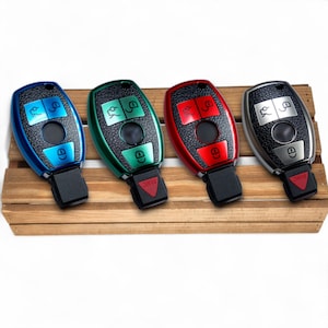 May include: Four car key fobs in blue, green, red, and silver. Each fob has a black center with buttons and a black base. The keys are arranged on a wooden surface. The buttons are in contrasting colors.
