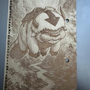 Peut inclure: Un cahier à spirales avec une illustration au bois brûlé détaillée d'Appa d'Avatar : Le dernier maître de l'air. L'image montre Appa volant au-dessus d'un paysage forestier. Le cahier est de couleur marron clair.