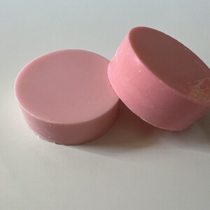 May include: Two round pink soap bars stacked on top of each other. The soap bars are smooth and have a slightly glossy finish.