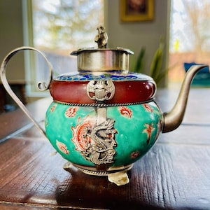 Pode incluir: Uma bule antigo e ornamentado, com uma alça e bico de prata. O bule apresenta um corpo turquesa com desenhos florais vermelhos e laranja e um dragão prateado. A tampa é prateada com uma pequena figura de macaco.