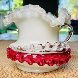 May include: A white glass pitcher with a ruffled rim and a ruby red ruffled base. The pitcher has a clear glass handle and a decorative, vintage design. The pitcher is likely used for serving beverages.
