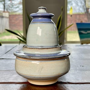 May include: A handmade ceramic jar with a lid. The jar is a light beige color with a blue band around the middle and top. The lid is a matching blue. The jar is sitting on a wooden table.