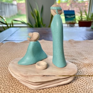 May include: A ceramic sculpture featuring two figures in teal robes with tan-colored heads and headbands. The figures stand on a light-colored, layered wooden base, with a small, round, tan-colored object in front of them. The sculpture is a decorative item.
