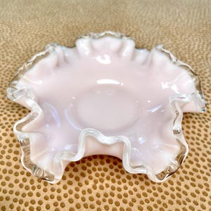 Puede incluir: Un cuenco de cristal rosa claro con borde con volantes y borde transparente e iridiscente. El cuenco tiene un centro poco profundo y redondeado y un borde delicado y ondulado. El cristal tiene una calidad suave y opalescente.