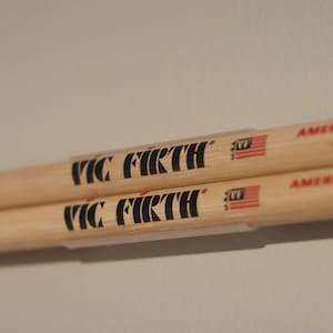 May include: Two Vic Firth American Classic 5A drumsticks. The drumsticks are made of light-colored wood and feature black text and a small American flag logo. The text "VIC FIRTH" is prominently displayed.