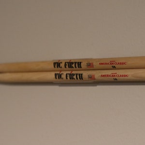 May include: Two wooden drumsticks, side by side, with the text "VIC FIRTH" in black. The drumsticks are labeled "AMERICAN CLASSIC 5B" in red. The drumsticks are made of light-colored wood.