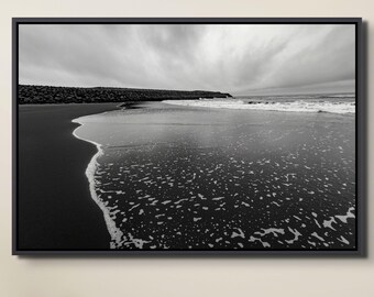 May include: Black and white framed photograph of a beach scene. The image captures the shoreline with dark sand and foamy waves. A cloudy sky fills the background, and a rock barrier is visible in the distance. The photograph is presented in a black frame.