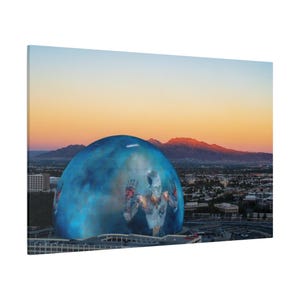 Könnte beinhalten: Eine große blaue Kugel mit einer darauf projizierten menschlichen Figur. Die Kugel befindet sich in einer Stadtlandschaft mit einem Gebirge im Hintergrund. Der Himmel ist in leuchtenden Orange- und Rosatönen.