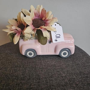 May include: A pink ceramic truck-shaped planter filled with artificial sunflowers. The truck has gray wheels and a white tag with the number 10. The sunflowers have cream and pink petals and brown centers.