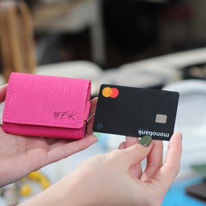 Könnte beinhalten: Eine leuchtend pinke Ledergeldbörse mit den Initialen "VFK" und einem Herzsymbol wird neben einer schwarzen Kreditkarte gezeigt. Die Kreditkarte hat das Mastercard-Logo und das Wort "monobank".