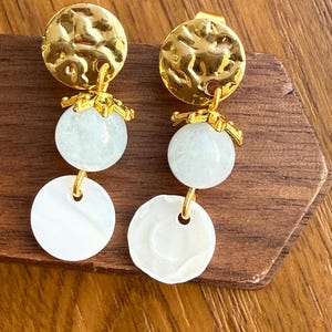 Puede incluir: Un par de pendientes dorados con textura martillada. Cada pendiente presenta un disco dorado, una esfera azul claro y un colgante blanco en forma de disco. Los pendientes se exhiben sobre una superficie de madera.