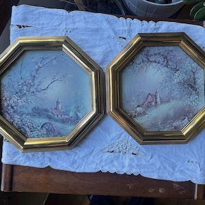 May include: Two octagonal wall art pieces featuring landscape paintings of rural farm scenes with barns, silos, and flowering trees. Each print is encased in a polished gold-toned frame with beveled edges, creating a matching set of decor.