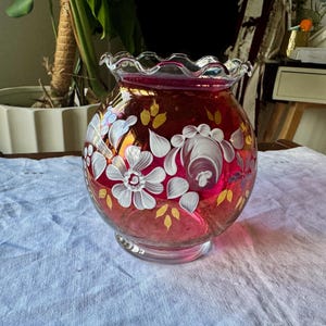 May include: A round, cranberry-colored glass vase with a ruffled, scalloped rim. The exterior features hand-painted white floral motifs and small yellow leaf accents. The glass is translucent, allowing light to pass through the deep red hue.