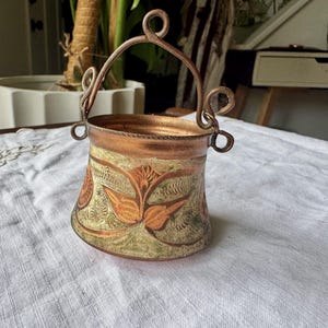 May include: A small copper bucket with a swinging handle and decorative scroll accents on the sides. The exterior features an etched floral pattern with green and orange patina highlights against the polished copper surface of the metal vessel.