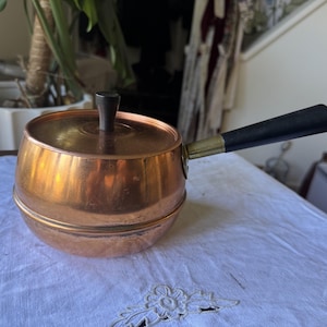 May include: A round copper pot with a matching lid and a long black handle. The lid features a small, cylindrical black knob at the center. The pot has a smooth, metallic finish with a horizontal seam running around the middle of the body.