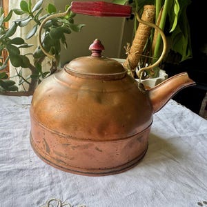 May include: A round copper tea kettle featuring a curved metal handle with a red wooden grip. The kettle has a matching lid with a small red wooden knob on top and a wide spout. The copper surface shows a natural patina with visible texture.