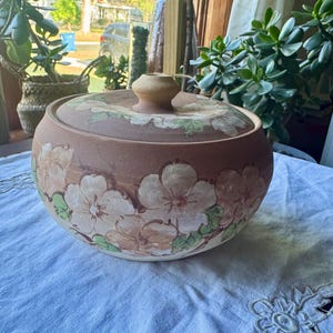 Pode incluir: Uma assadeira redonda de cerâmica na cor terracota com tampa a condizer. O exterior apresenta um padrão floral pintado à mão em tons de creme, rosa claro e verde. A tampa tem um puxador central e um design floral correspondente.