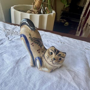 May include: A ceramic cat figurine with a beige base, featuring hand-painted blue stripes along the back and tail, and intricate green leaf patterns. The cat has a black-tipped tail, dark facial markings, and a rounded, stylized body shape.