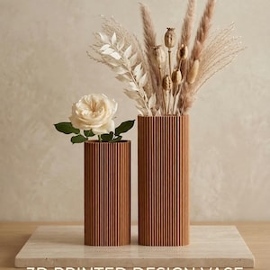 Peut inclure: Deux vases marron imprimés en 3D avec des rainures, l'un contenant une rose blanche et l'autre des fleurs séchées. Les vases sont posés sur une surface semblable à du marbre. Le texte en bas indique "3D PRINTED DESIGN VASE" avec "14 COULEURS | 3 TAILLES".