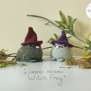 Może przedstawiać: Dwie pluszowe zabawki w kształcie żaby czarownicy, wykonane na szydełku, z czapkami w szpic. Jedna żaba ma czapkę bordową, a druga fioletową. Żaby siedzą na białym tle, na którym leży zielony mech i gałązka.