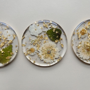 May include: Three clear resin coasters with dried flowers and gold leaf inclusions. Each coaster features a different arrangement of white and yellow flowers and green leaves, creating a natural, decorative look.