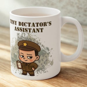 以下が含まれることがあります： 「TINY DICTATOR'S ASSISTANT」の文字が茶色でプリントされた白いセラミックマグ。マグには、軍服を着てクリップボードを持っている人の漫画イラストが描かれています。背景は灰色のスプラッターデザインです。