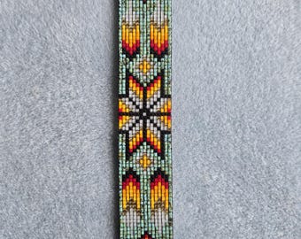 May include: A beaded bookmark with a geometric pattern in shades of orange, yellow, red, and turquoise. The design includes star and arrow shapes, with black thread at the top and bottom. The bookmark is approximately 2.5 cm wide.