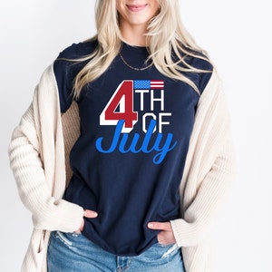 以下が含まれることがあります： アメリカの国旗と赤い「4th of July」のグラフィックがプリントされたネイビーのTシャツを着た女性。彼女は青いジーンズとクリーム色のカーディガンも着ています。