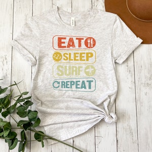 May include: A light gray t-shirt with a vintage-style graphic that reads "Eat, Sleep, Surf, Repeat". The graphic is in a rainbow of colors with a surfboard, a sleeping face, and a fork and knife.