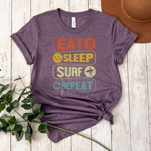 May include: Purple t-shirt with a vintage-style graphic that reads "Eat, Sleep, Surf, Repeat". The graphic is in a rainbow of colors with a surfer icon.
