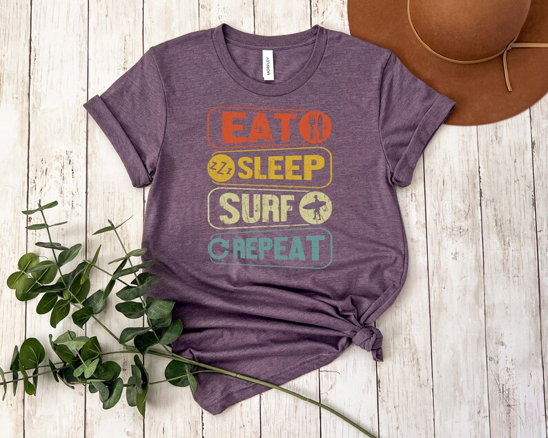 May include: Purple t-shirt with a vintage-style graphic that reads "Eat, Sleep, Surf, Repeat". The graphic is in a rainbow of colors with a surfer icon.