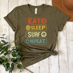 May include: Olive green t-shirt with a vintage-style graphic that reads "Eat, Sleep, Surf, Repeat". The graphic is in a rainbow of colors with a surfboard icon.