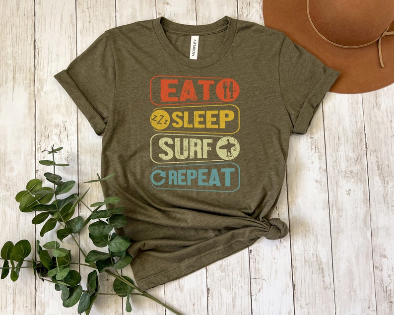 May include: Olive green t-shirt with a vintage-style graphic that reads "Eat, Sleep, Surf, Repeat". The graphic is in a rainbow of colors with a surfboard icon.