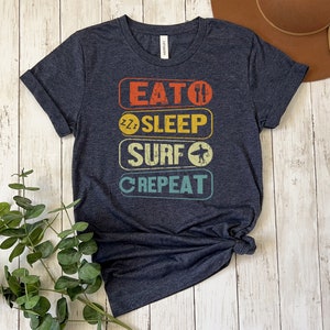 May include: A dark blue t-shirt with a vintage-style graphic that reads "Eat, Sleep, Surf, Repeat". The graphic is in a rainbow color scheme.