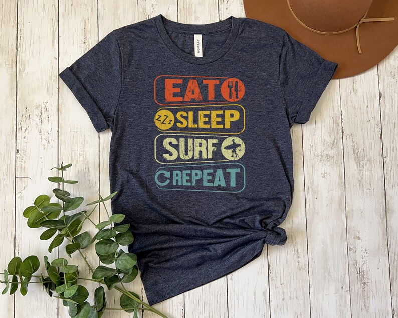 May include: A dark blue t-shirt with a vintage-style graphic that reads "Eat, Sleep, Surf, Repeat". The graphic is in a rainbow color scheme.