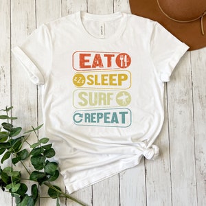 May include: White t-shirt with a vintage-style graphic that reads "Eat, Sleep, Surf, Repeat". The graphic is in a rainbow color scheme with a fork and knife, a sleeping face, a surfboard, and a circular arrow.