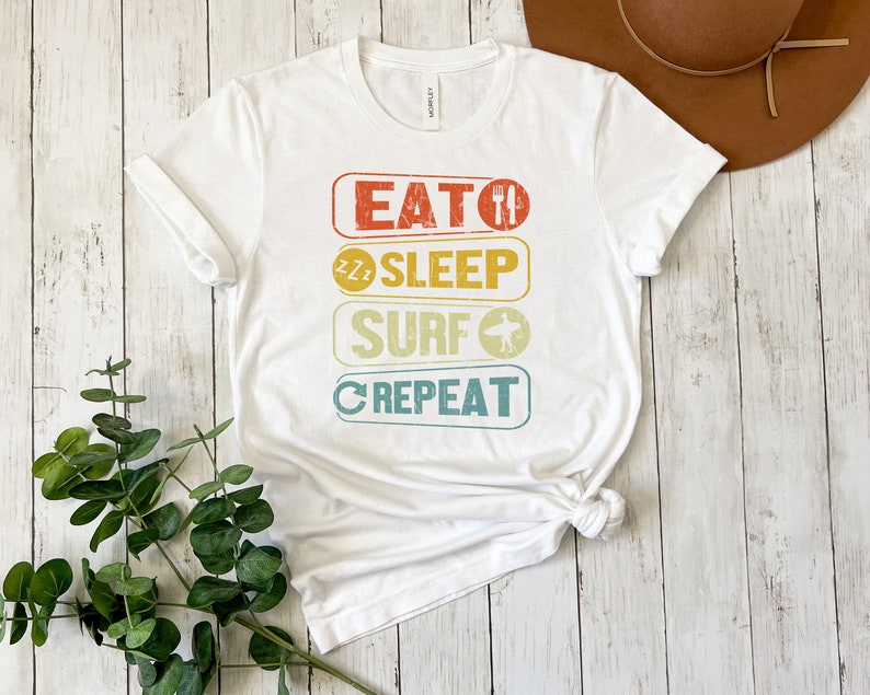 May include: White t-shirt with a vintage-style graphic that reads "Eat, Sleep, Surf, Repeat". The graphic is in a rainbow color scheme with a fork and knife, a sleeping face, a surfboard, and a circular arrow.