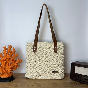 May include: A white crocheted tote bag with brown leather straps and a small brown leather label that says "SKH".