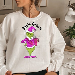 May include: White sweatshirt with a green Grinch character wearing a pink sparkly Santa hat and holding a pink sparkly cup. The text "Basic Grinch" is above the character.